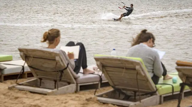 Deux vacanciers européens profitent de chaises longues sur une plage du Sahara occidental tandis qu'un autre pratique le kitesurf dans les vagues